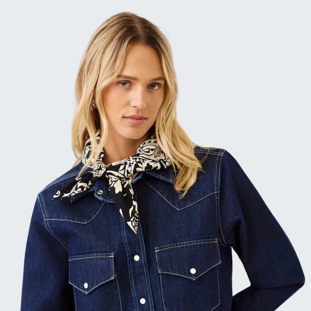 A woman wearing a dark indigo denim western-style shirt jacket with silver snap buttons and chest pockets, styled with a black and white patterned neck scarf, against a light gray studio background.