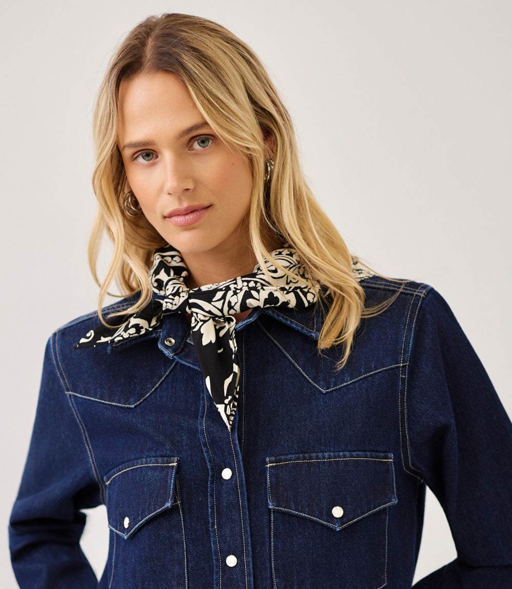 A woman wearing a dark blue denim western-style jacket with a black and white patterned bandana tied around her neck, photographed against a light gray studio background.