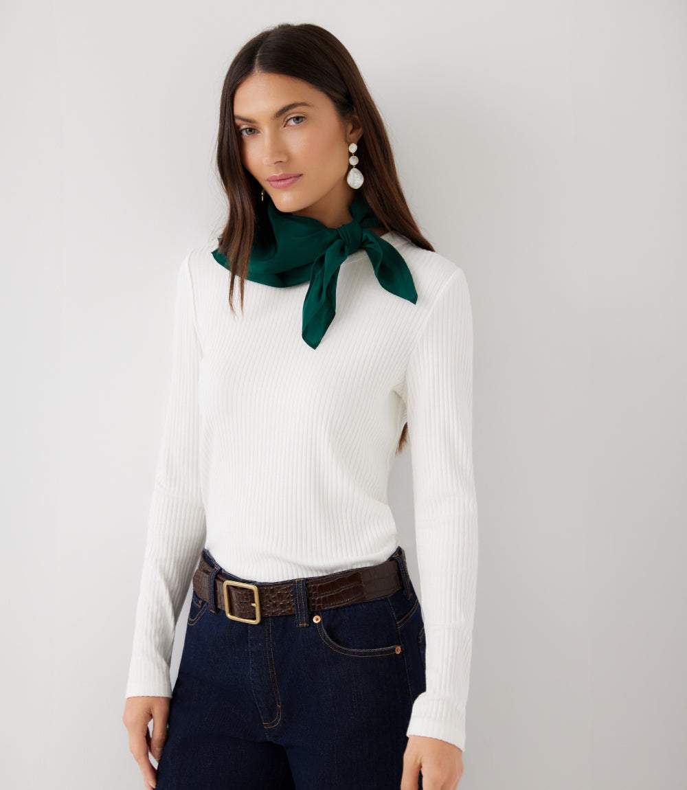 A woman modeling a white ribbed long-sleeve top with a dark green silk neck scarf tied at the collar, paired with dark blue jeans and a brown belt, against a light gray studio background.