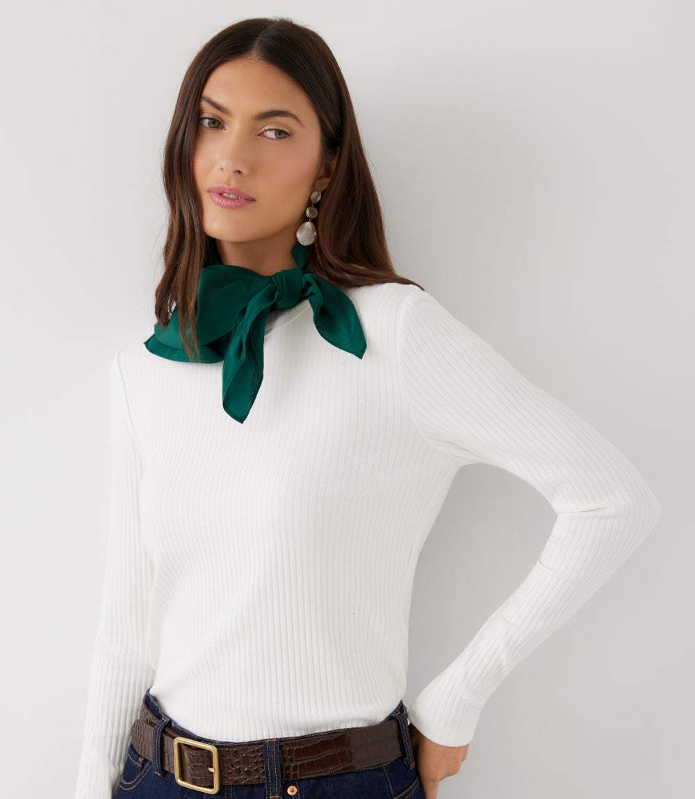 A woman wearing a white ribbed long-sleeve top with a dark green satin neck bow, paired with dark blue jeans and a brown belt, against a light gray studio background.