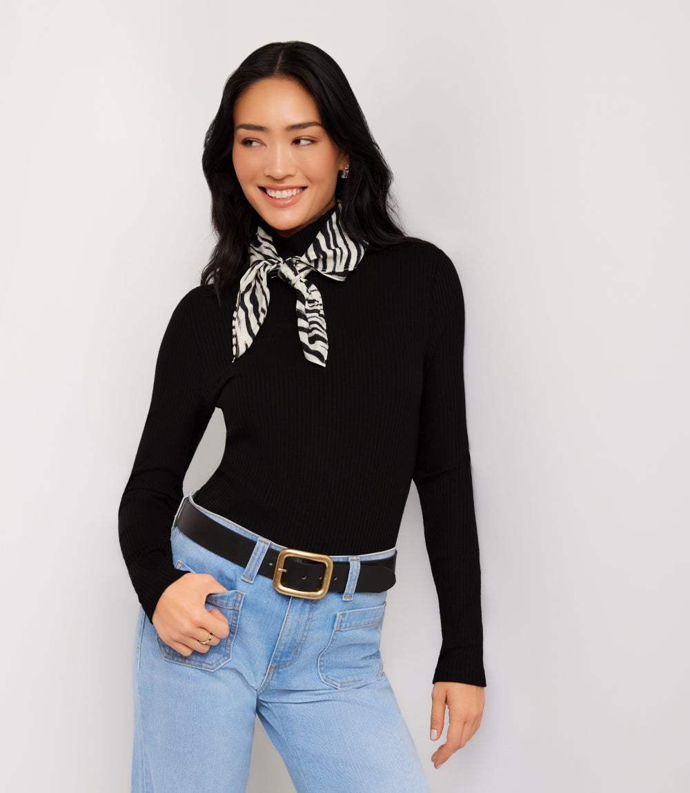A woman modeling a black ribbed long-sleeve turtleneck sweater with a black and white zebra-print neck scarf, paired with light blue jeans and a black belt, against a light gray studio background.