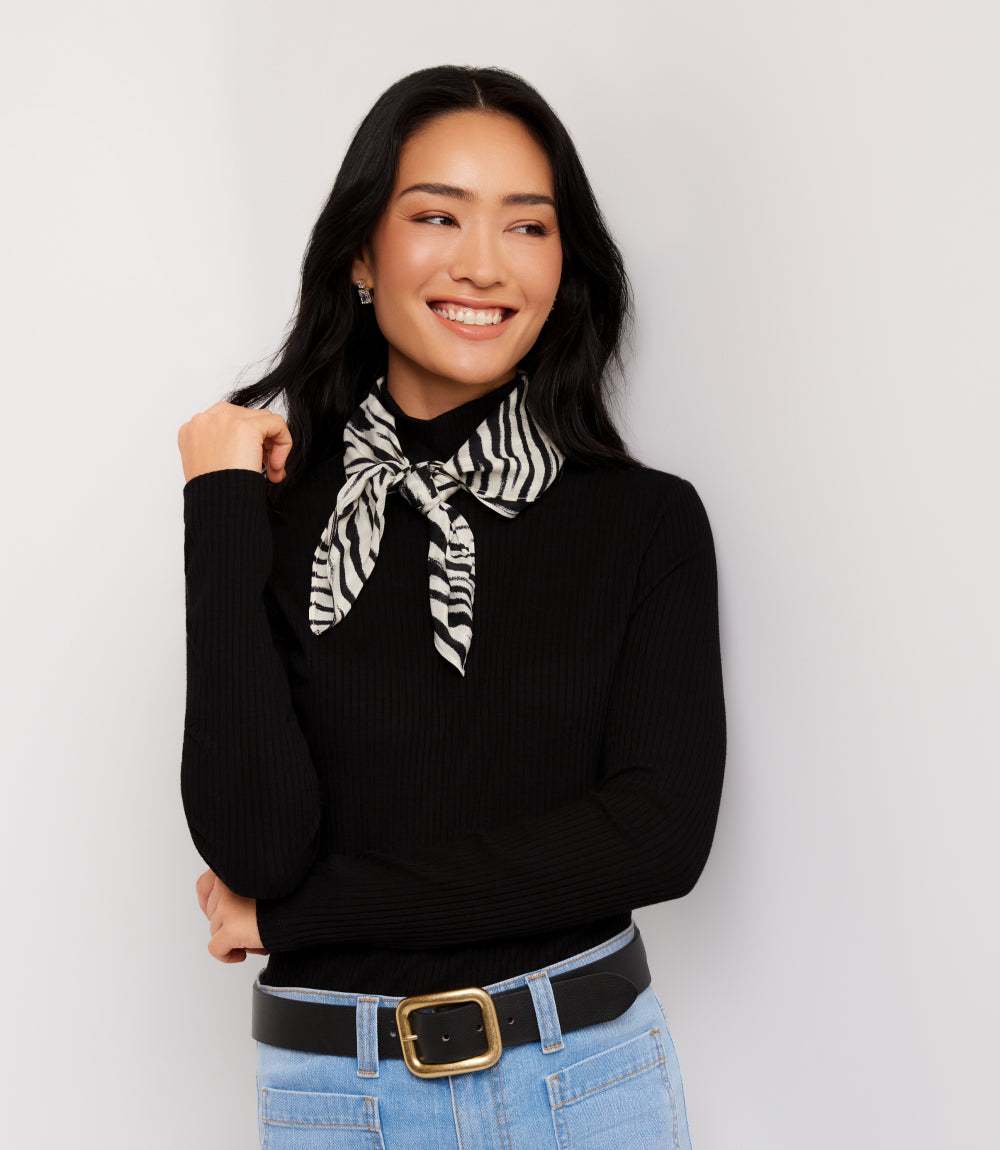 A woman wearing a black ribbed long-sleeve turtleneck sweater with a black and white zebra-print neck scarf tied at the collar, paired with light blue jeans and a black belt with a gold buckle, against a plain light gray background.
