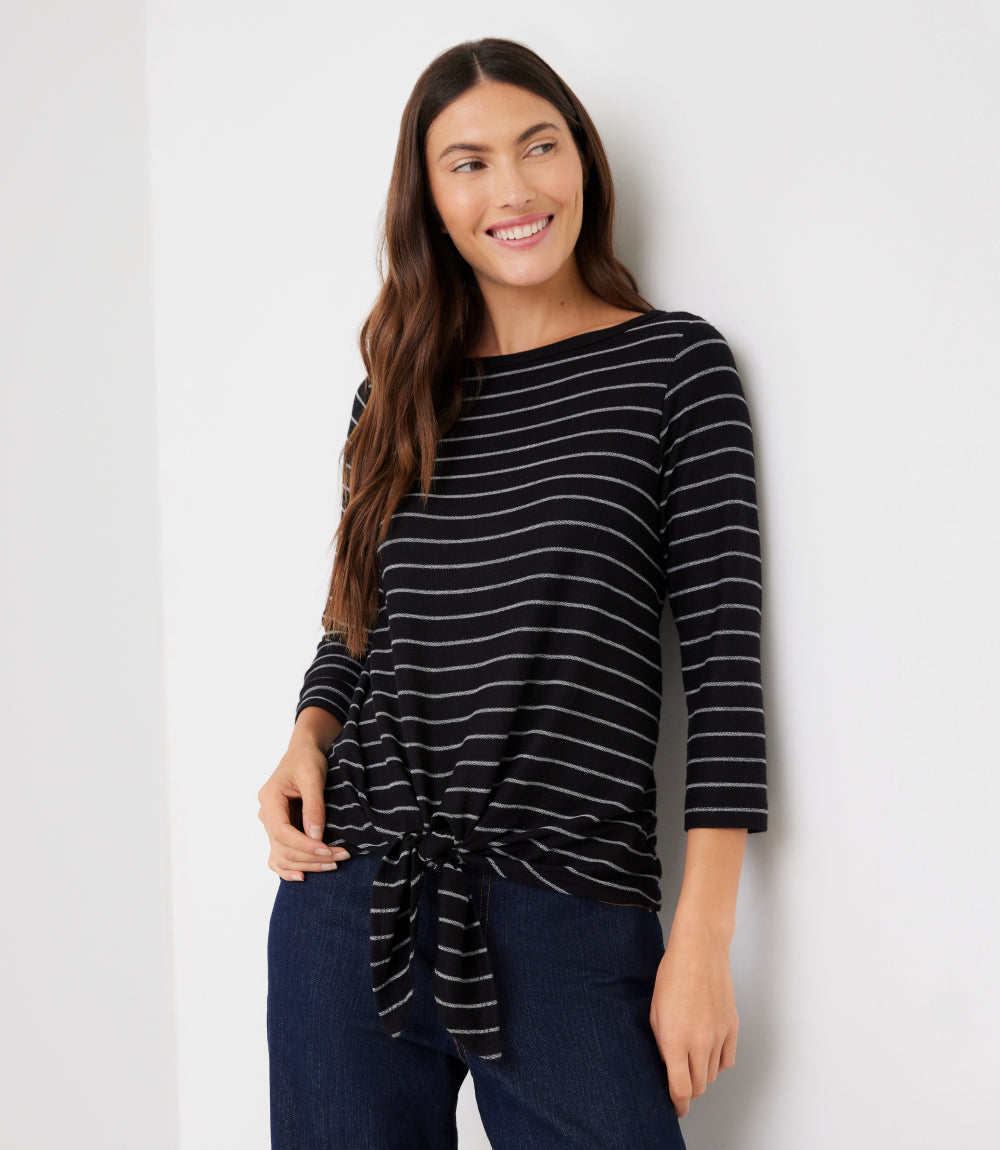 A woman wearing a black and white striped three-quarter sleeve top with a front knot detail, paired with dark blue jeans, standing against a plain white wall.