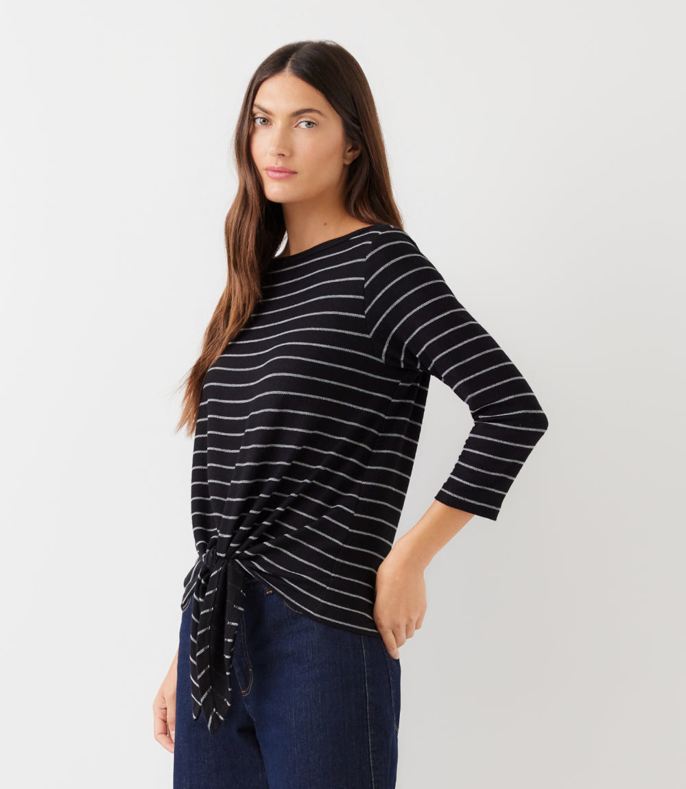 A woman modeling a black and white striped knit top with three-quarter sleeves and a side tie detail, paired with dark blue jeans against a plain light gray background.
