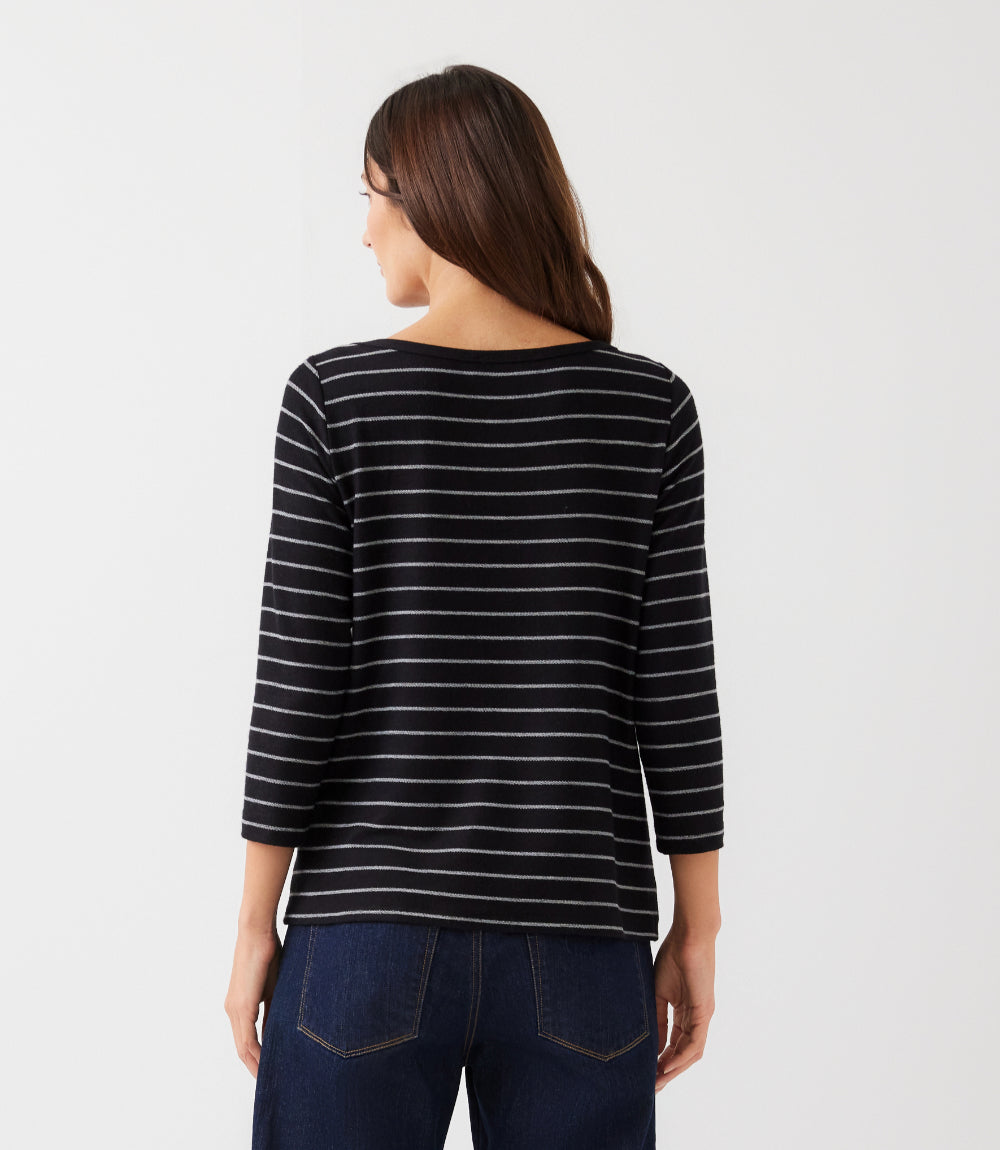 A woman viewed from the back wearing a black three-quarter sleeve top with thin white horizontal stripes and dark blue jeans, set against a plain light gray studio background.