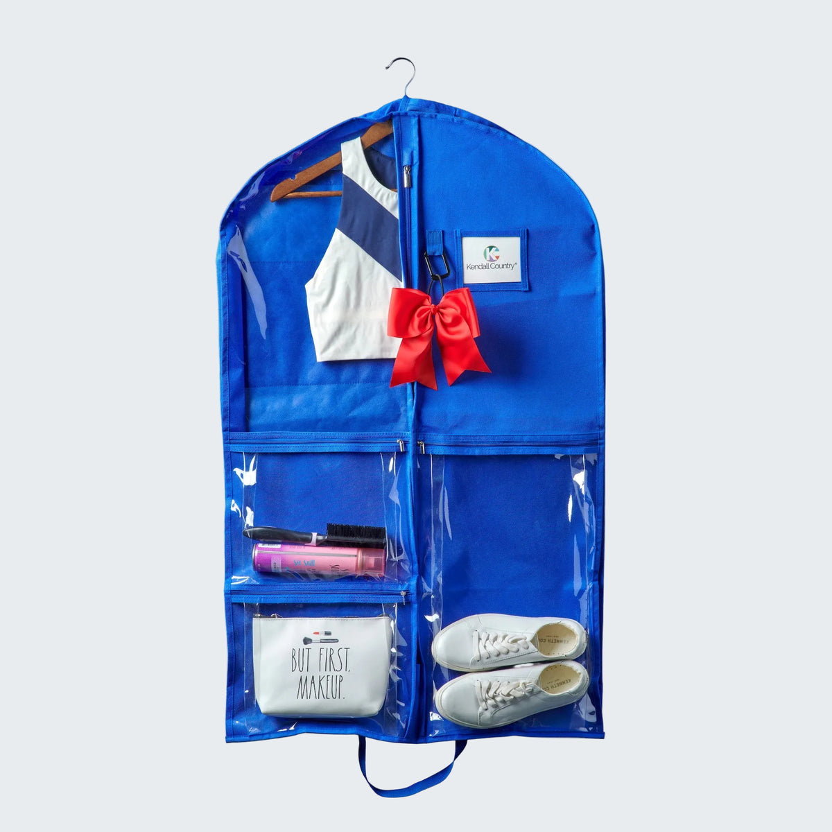 Blue garment bag with clear front pocket containing a white shirt, red bow, shoes, and makeup items on a white background.