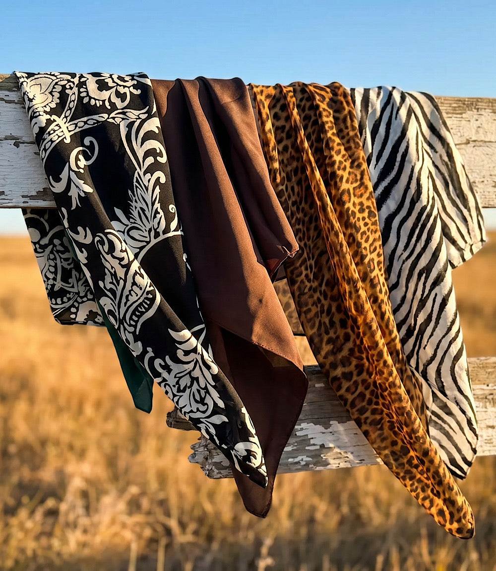 Four patterned fabric scarves or wraps draped over a weathered wooden fence outdoors, with a field and blue sky in the background. The fabrics show black-and-white damask, solid brown, leopard print, and zebra print patterns.