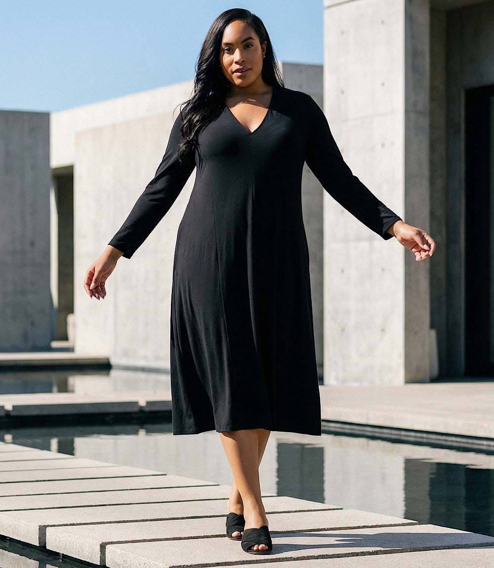 A woman wearing a black long-sleeve midi dress walks across a modern concrete walkway next to a reflecting pool.