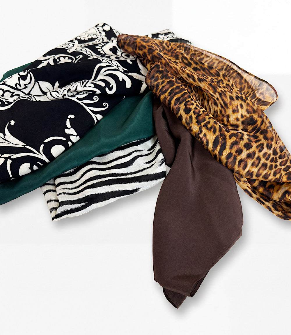 A pile of folded patterned scarves on a light gray background, including black and white damask, teal green, black and white zebra, brown, and leopard print.
