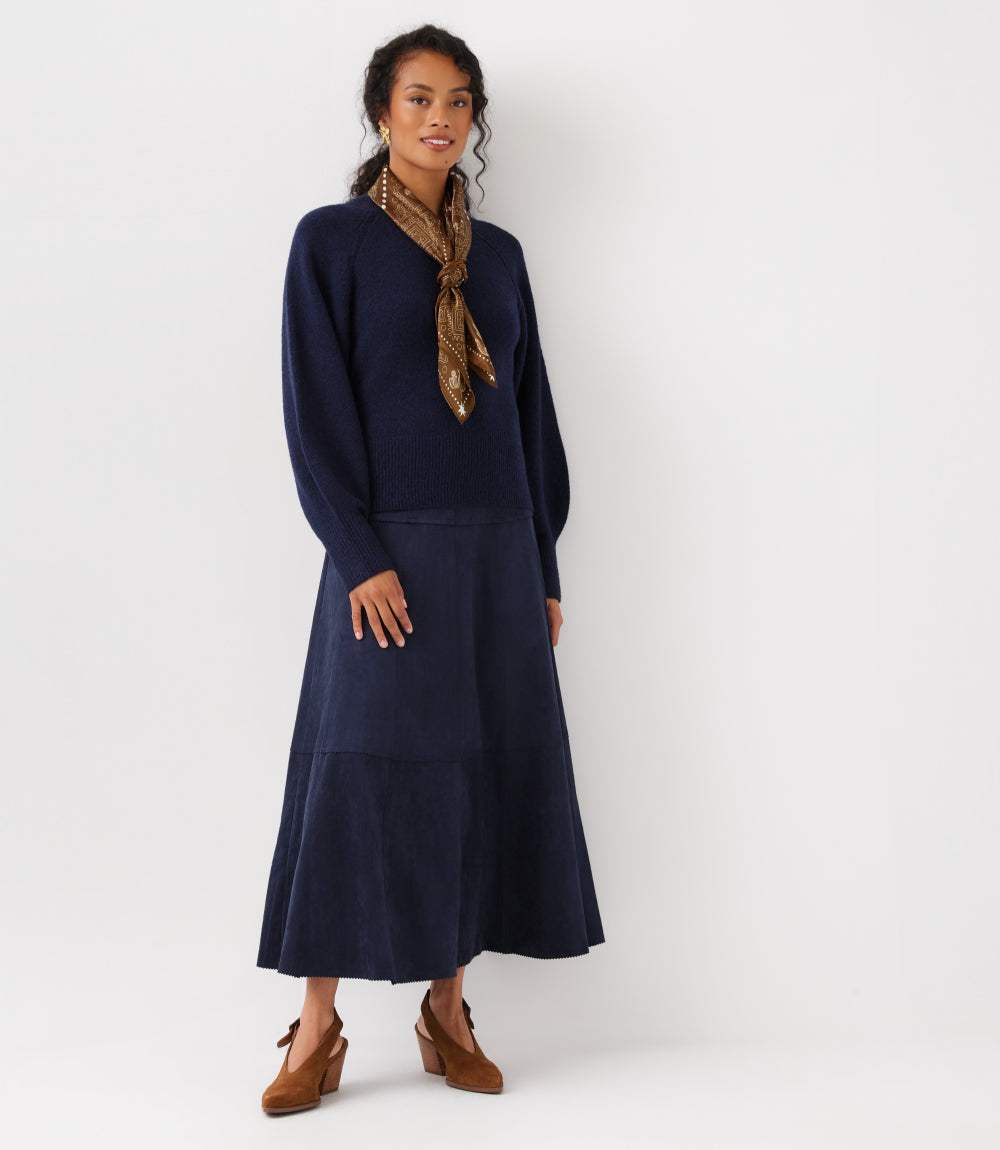 A woman modeling a navy blue long-sleeve midi dress with a tie-neck scarf and brown suede shoes against a plain white studio background.
