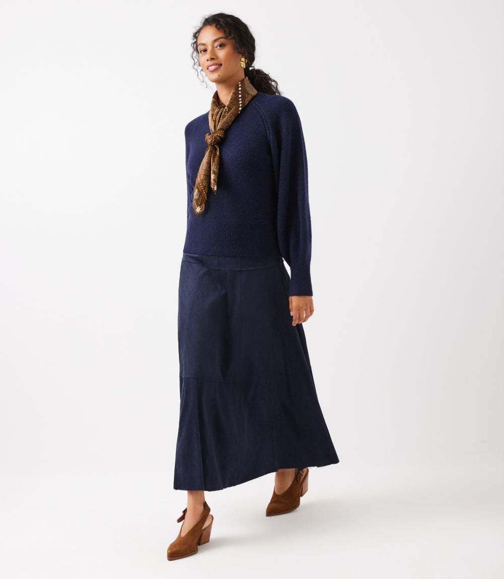 A woman modeling a navy blue knit sweater and matching midi skirt set, styled with a brown patterned scarf and brown shoes, against a plain white studio background.