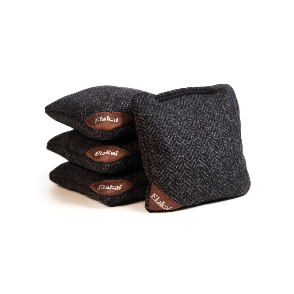 Set of three dark gray herringbone patterned cornhole bags with brown leather patches on a white background.