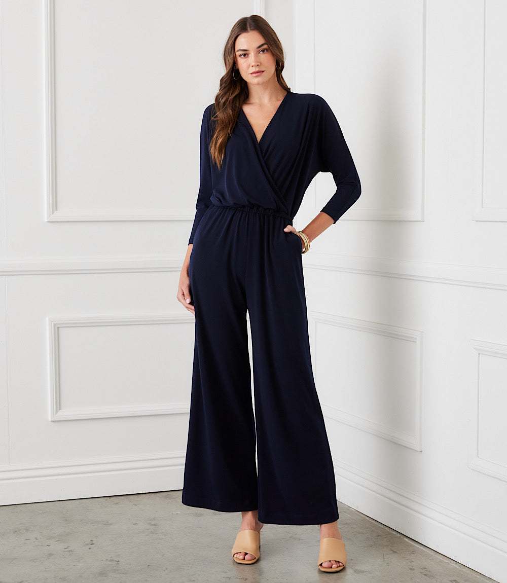 A woman modeling a navy blue wrap-style jumpsuit with wide legs and elbow-length sleeves, standing against a white paneled wall.