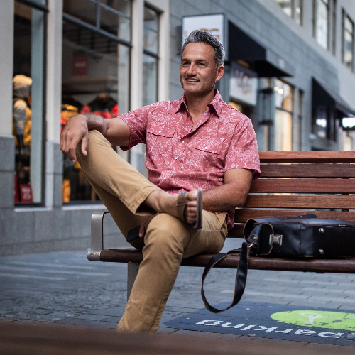 Man sitting on a bench wearing a pink shirt and beige pants in an urban setting.