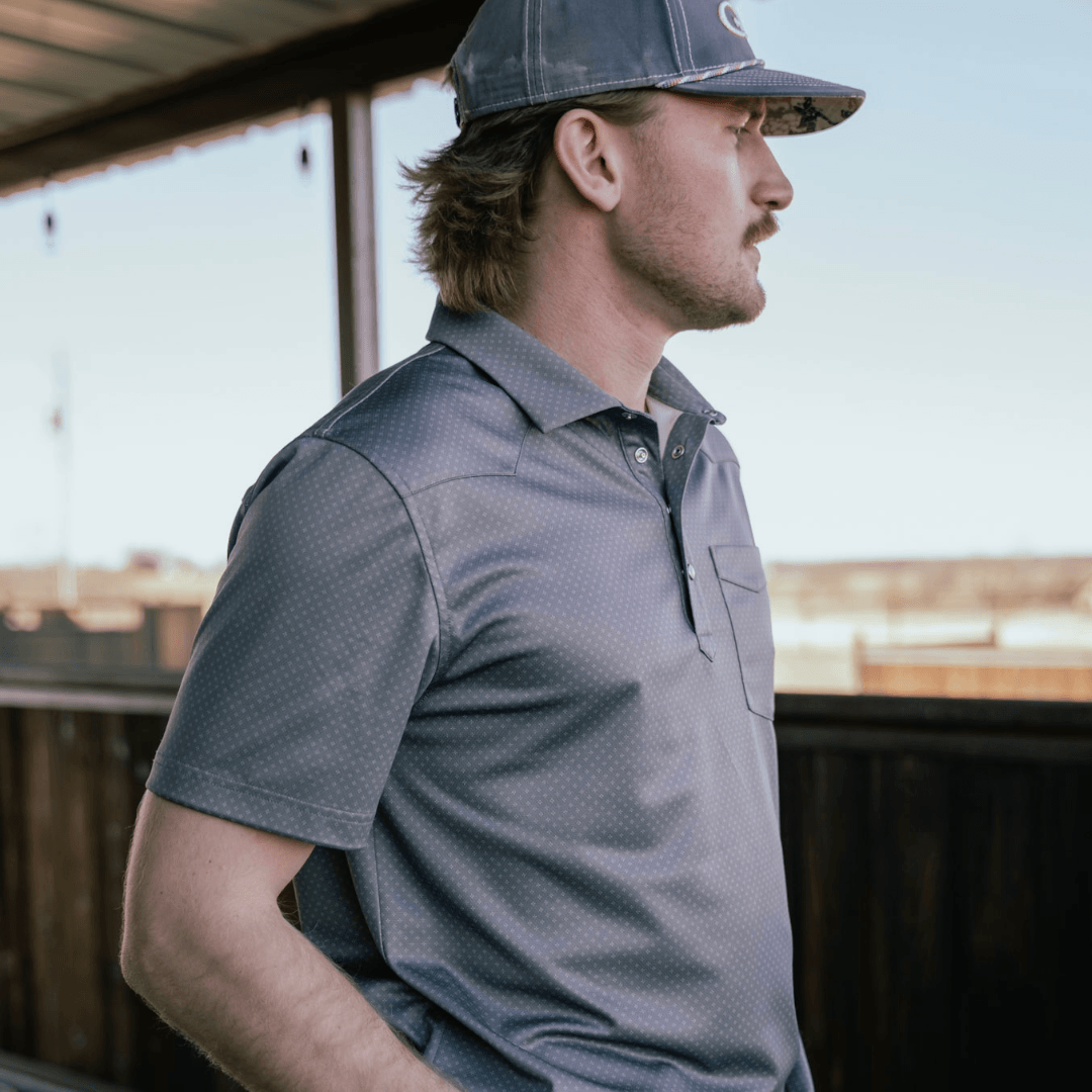 A man in profile wearing a dark gray short-sleeve polo shirt and a matching dark gray baseball cap, standing on an outdoor porch.