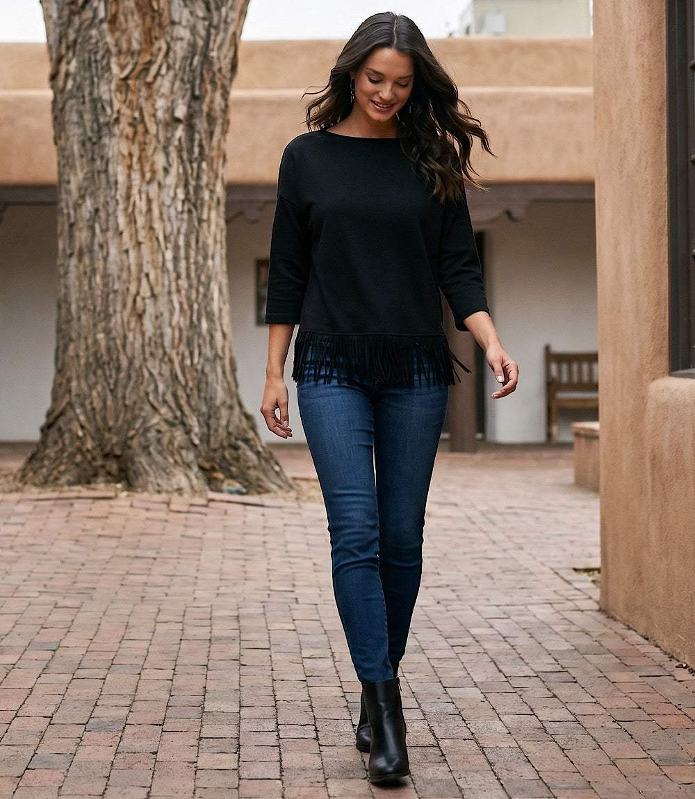 A woman walking outdoors on a brick patio wearing a black long-sleeve top with fringe trim, dark blue skinny jeans, and black ankle boots.