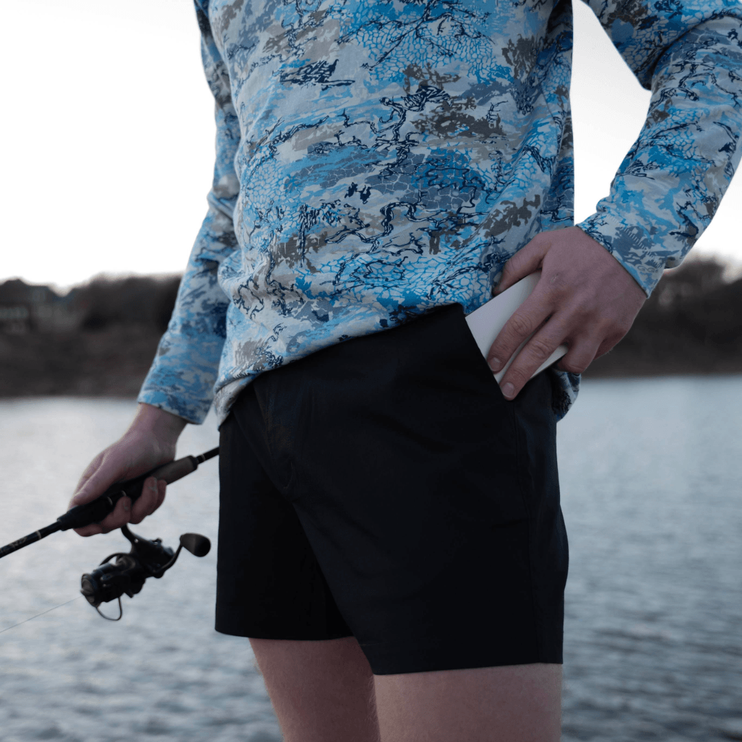 Person wearing a blue camouflage jacket and black shorts, holding a fishing rod by a body of water.
