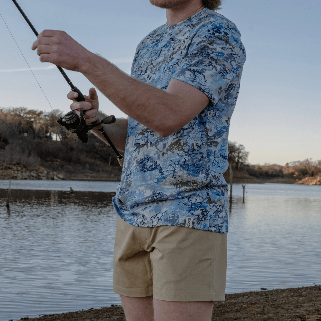 Person fishing by a lake wearing a blue camouflage shirt and beige shorts.