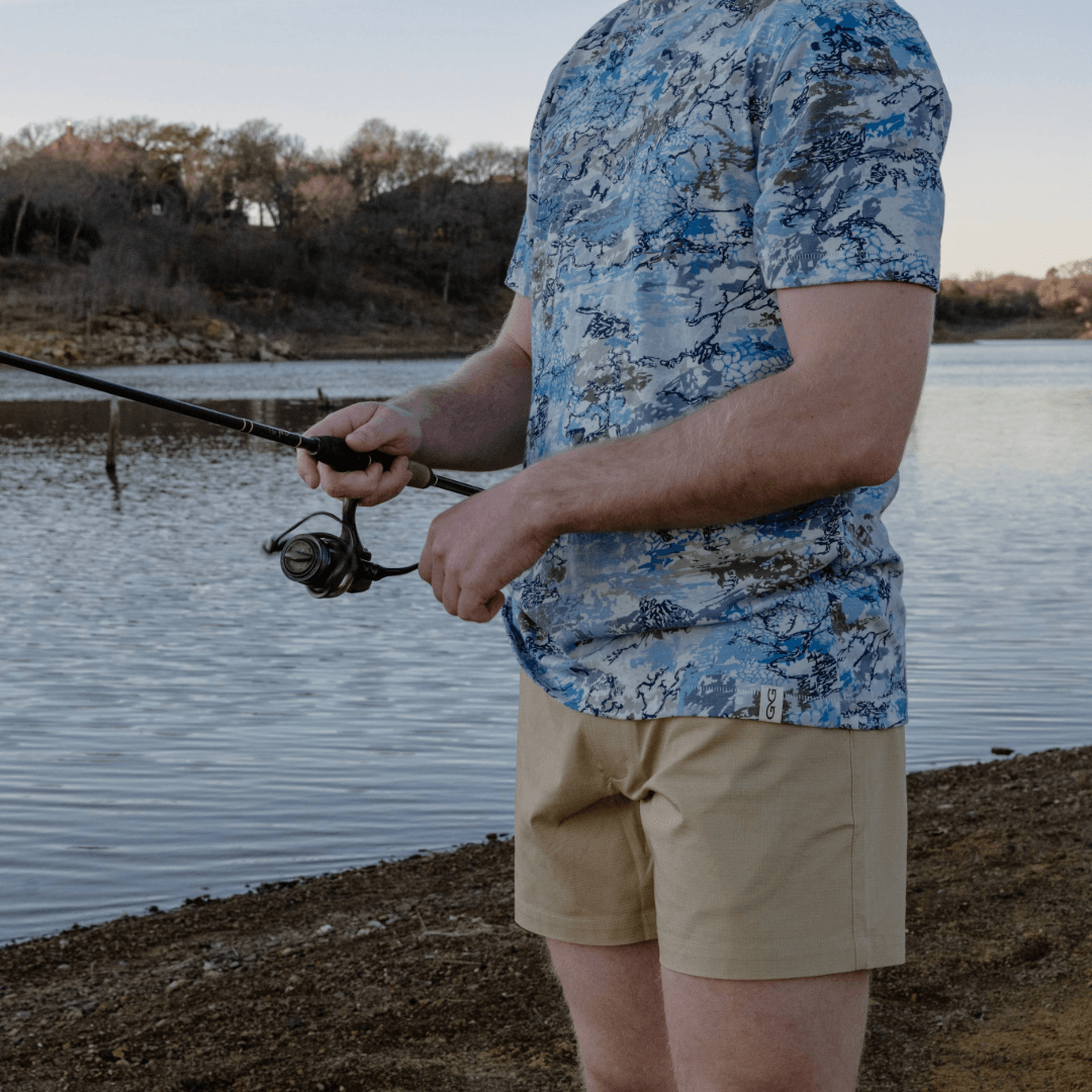 Person fishing by a lake wearing a blue camouflage shirt and beige shorts.
