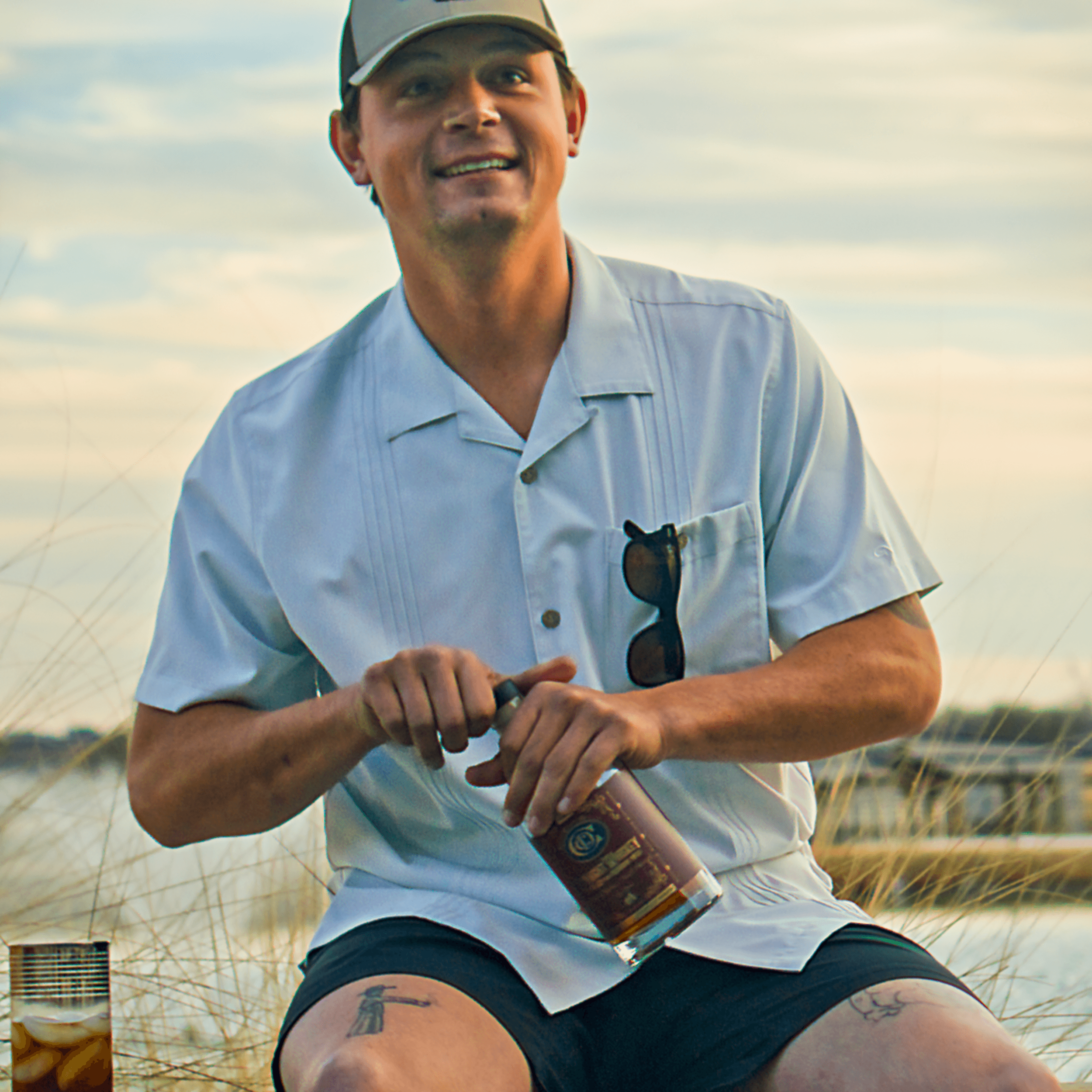 Man holding a bottle of whiskey on a beach with a sunset in the background