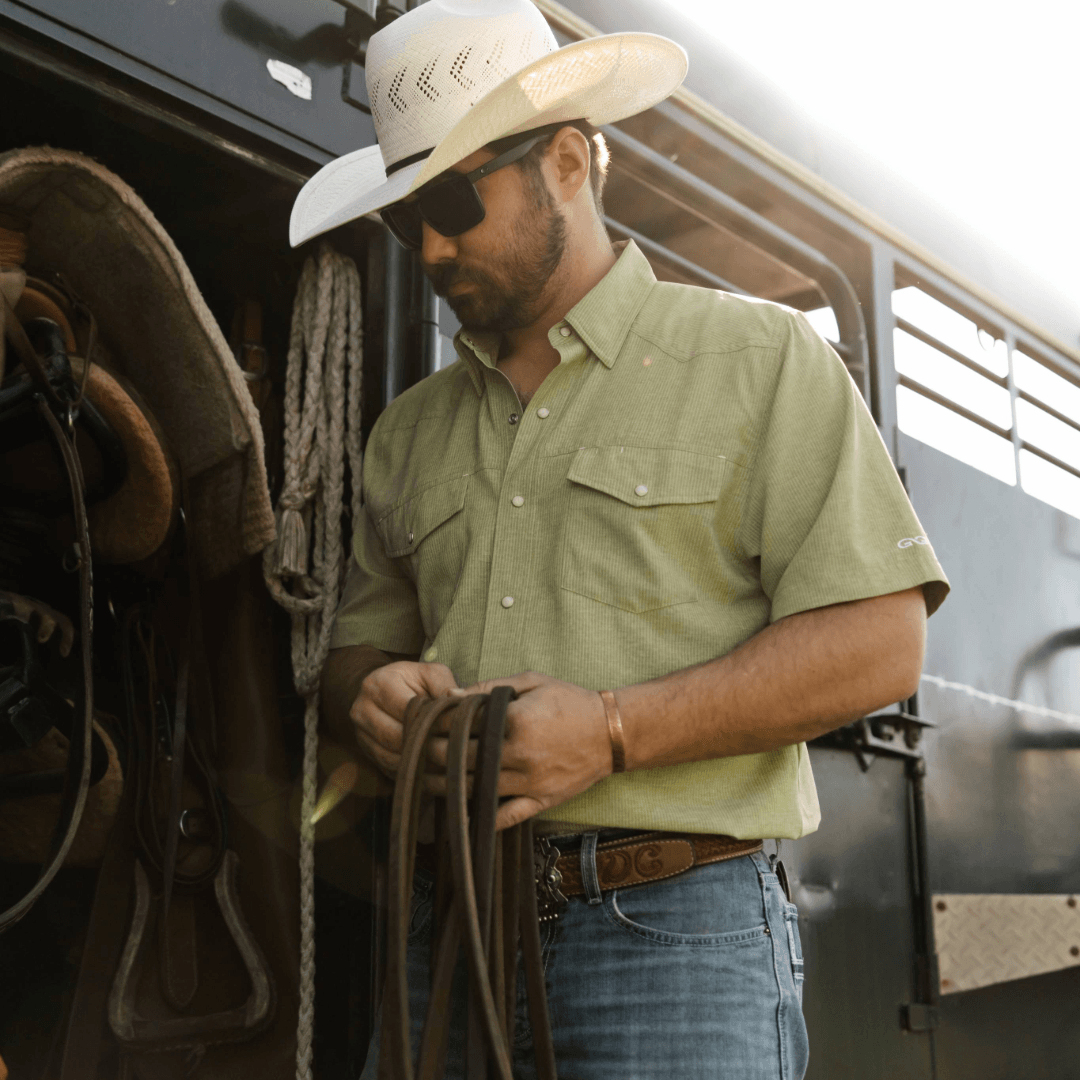 Man in a cowboy hat and green shirt holding a rope next to horse tack