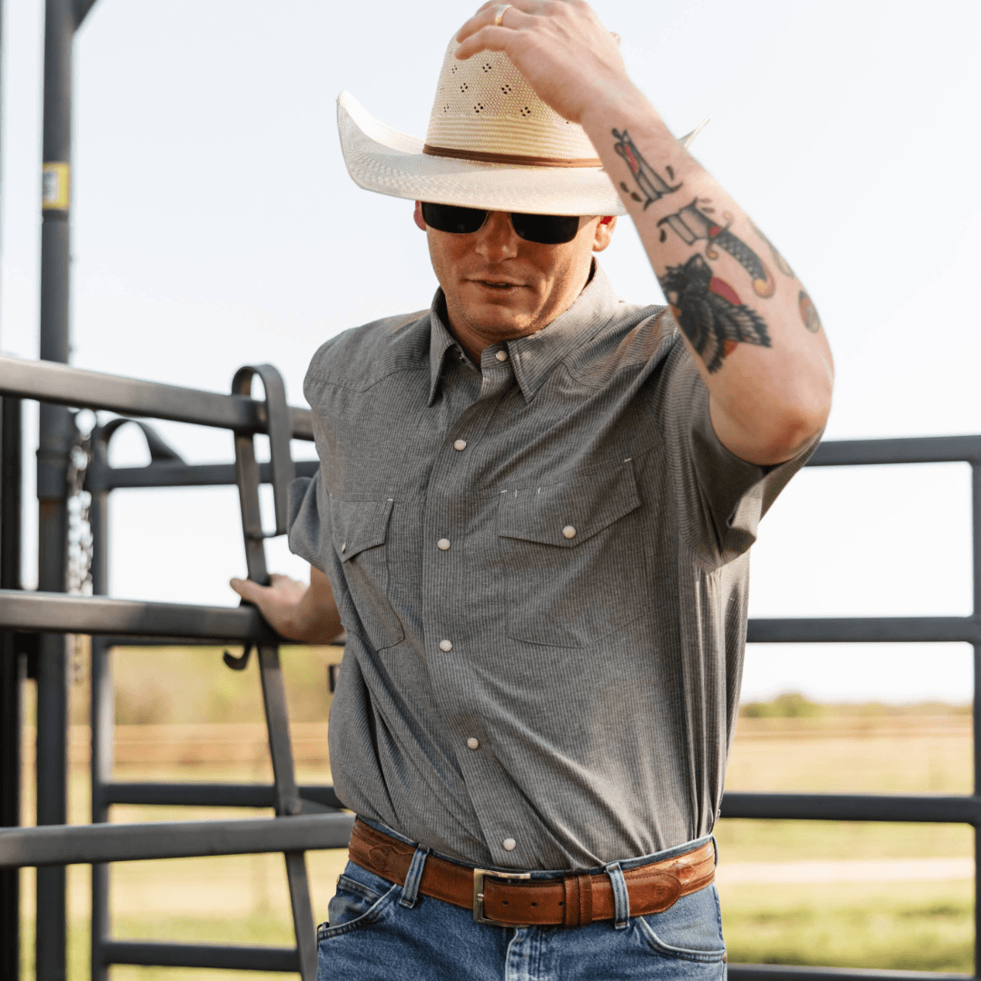 Man wearing a cowboy hat and sunglasses, standing in front of a metal gate with a rural landscape in the background.