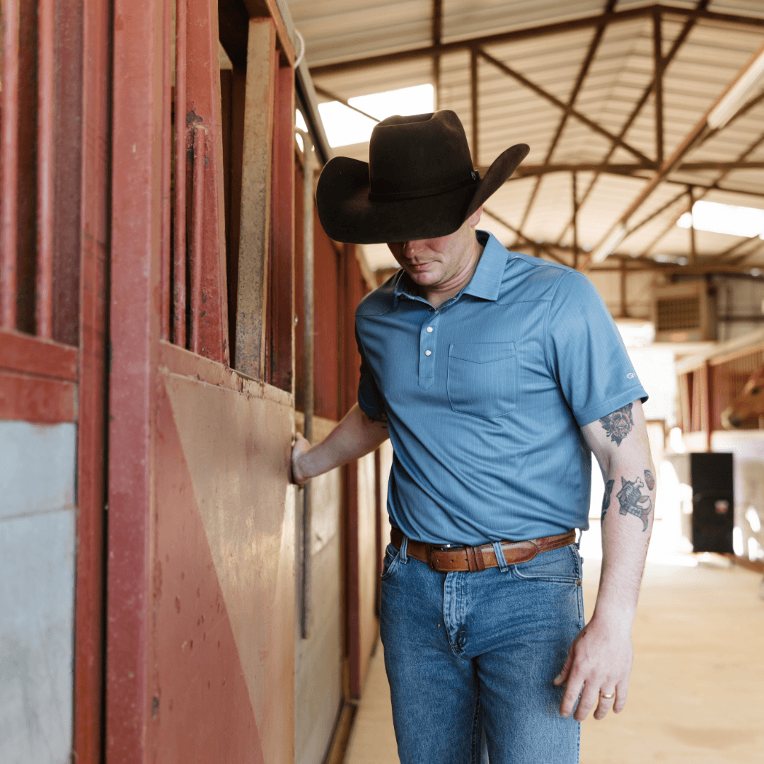 Man in a blue shirt and cowboy hat standing next to a red barn door.