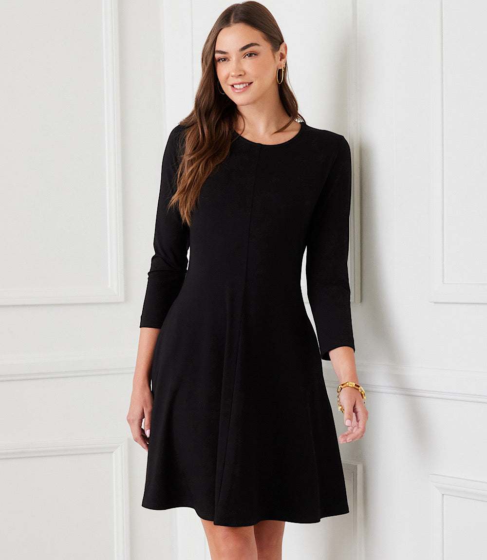 A woman modeling a black three-quarter sleeve fit-and-flare dress against a white paneled wall.