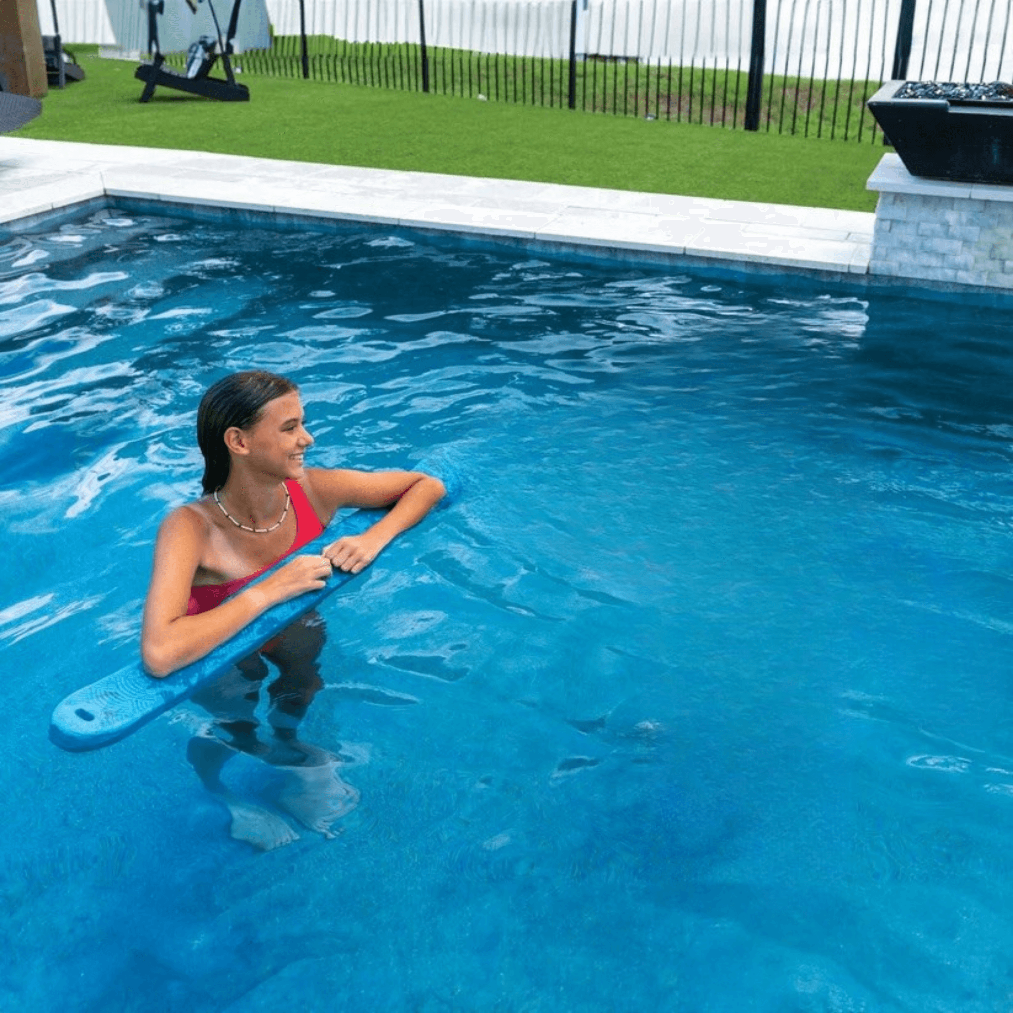 Woman in a pool holding a blue pool noodle with grass and a fence in the background