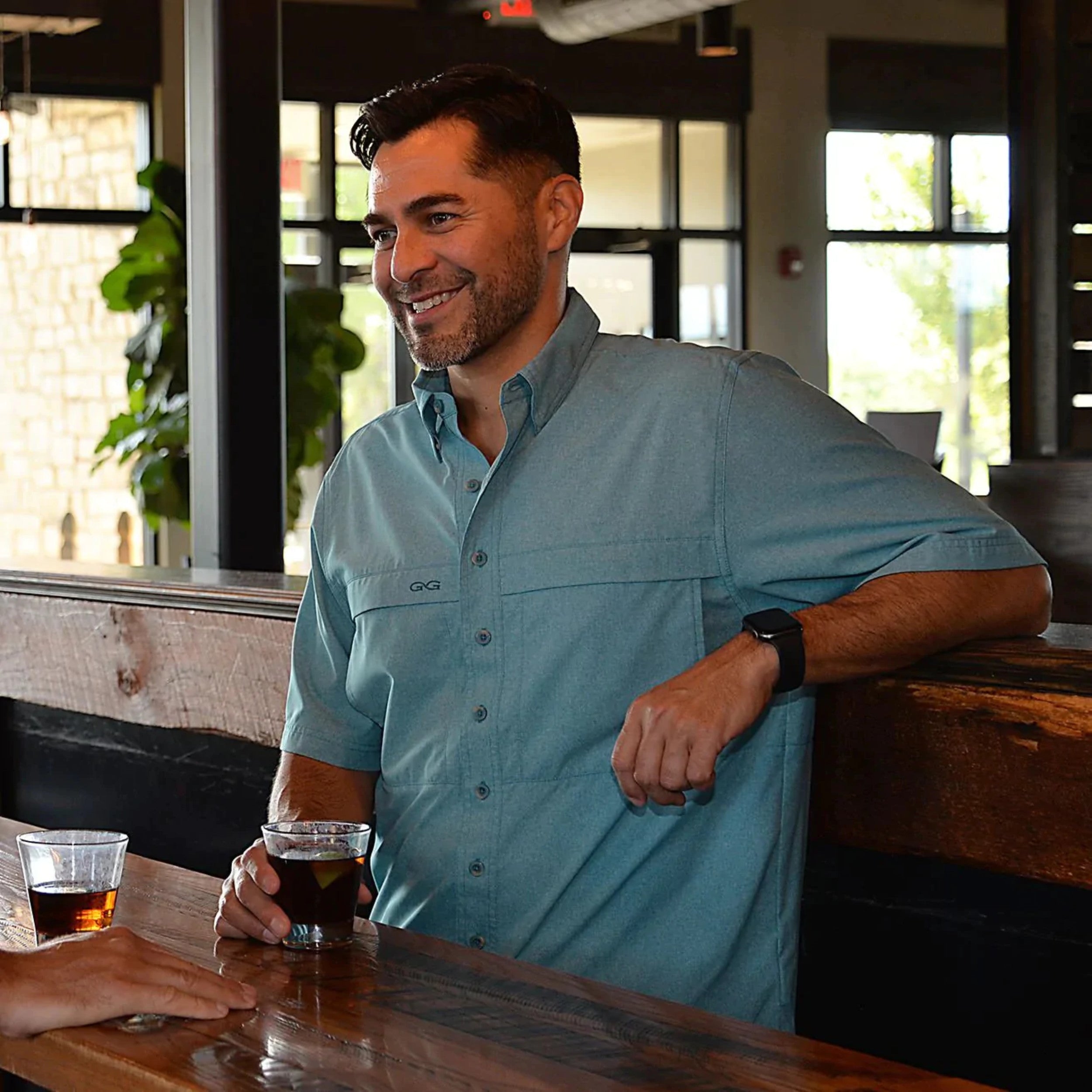 Man in a blue shirt sitting at a bar with drinks, smiling.