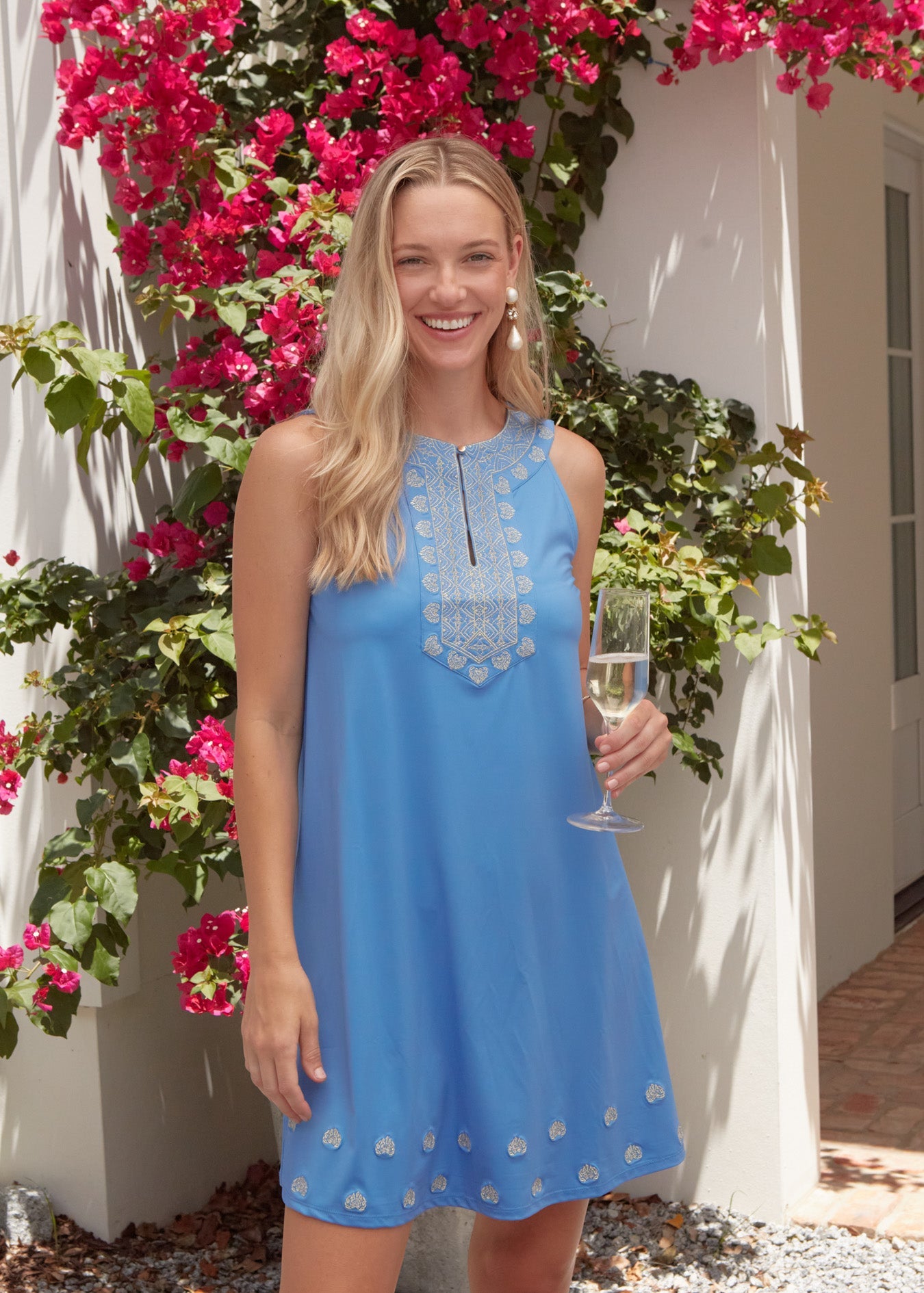 Woman in a blue dress holding a glass of wine in front of pink flowers.