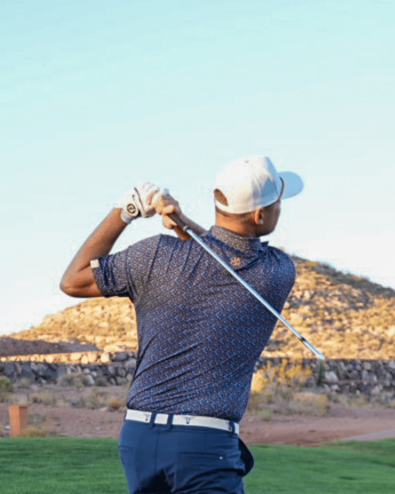Golfer in a blue shirt and white cap swinging a golf club with mountains in the background