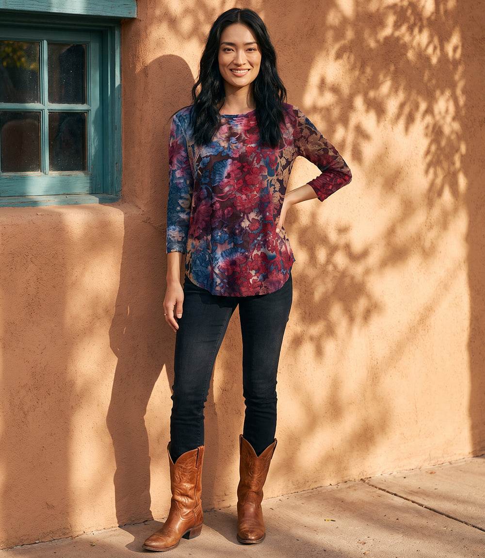 A woman stands outdoors against a warm stucco wall with tree shadows, wearing a long-sleeve floral top with blue, purple, and red tones, black skinny jeans, and brown cowboy boots.