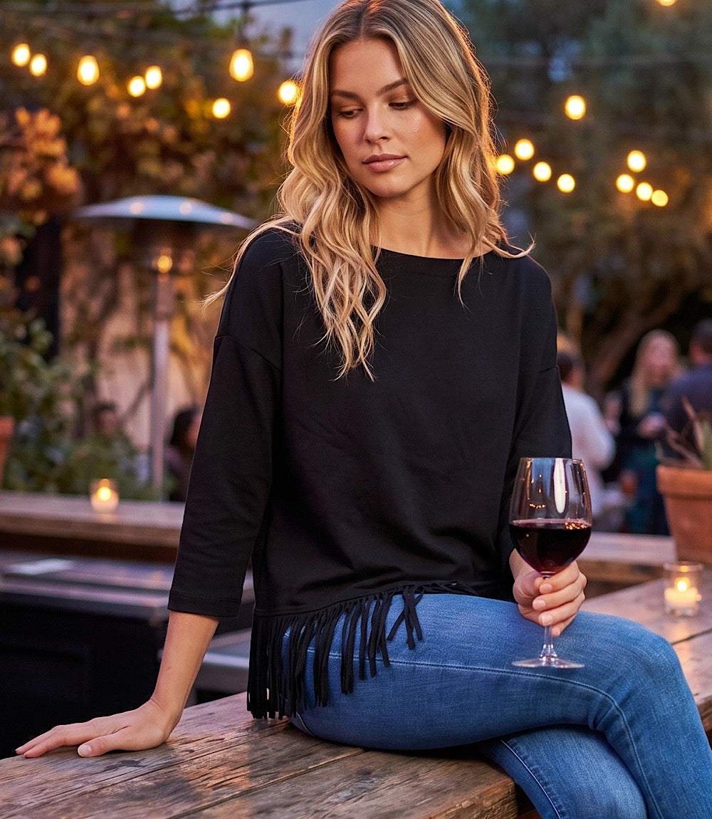 A woman sitting on a wooden table at an outdoor patio, wearing a black three-quarter sleeve top with fringe trim and blue jeans, holding a glass of red wine.