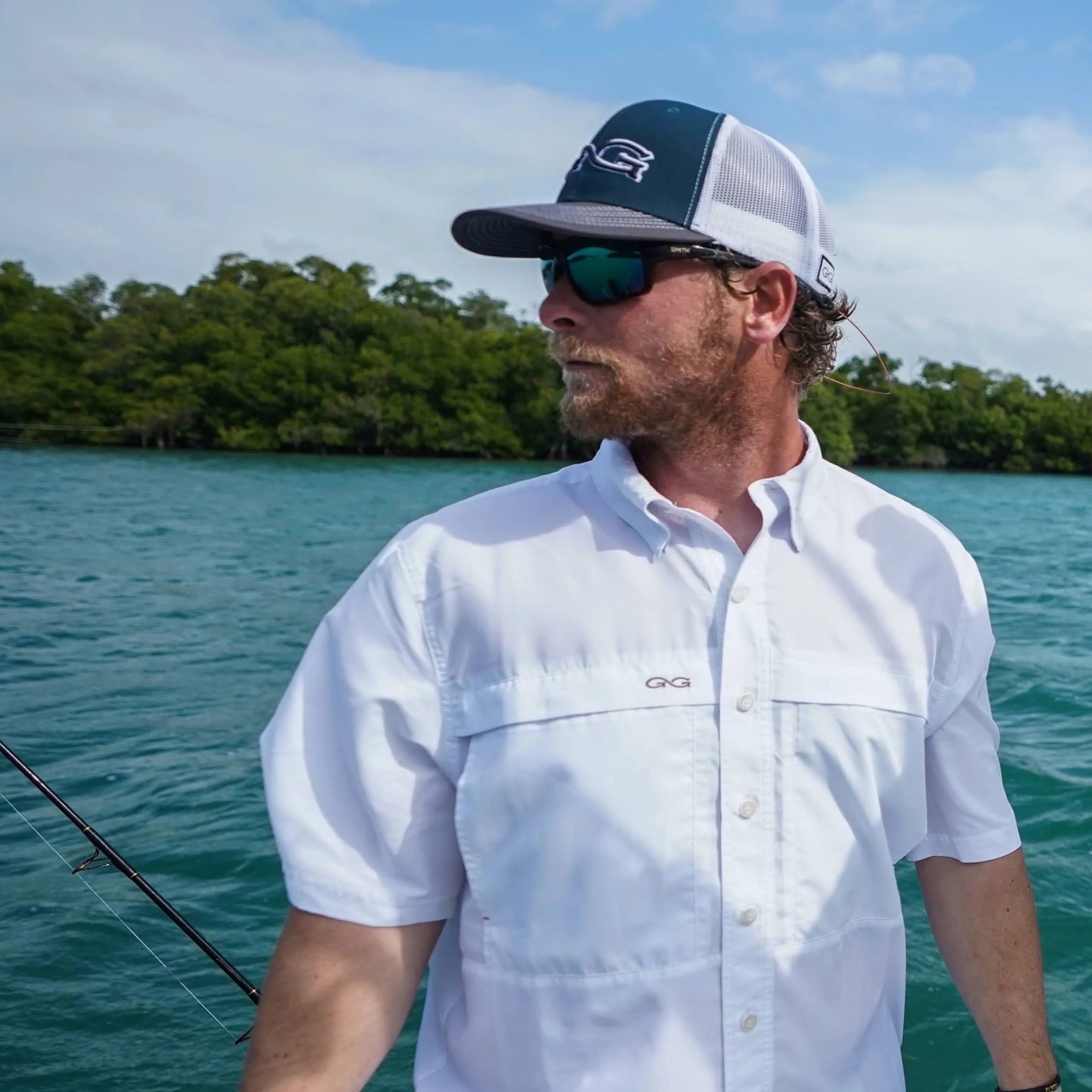 Man wearing a white shirt and cap with sunglasses, standing on a boat with water and trees in the background.