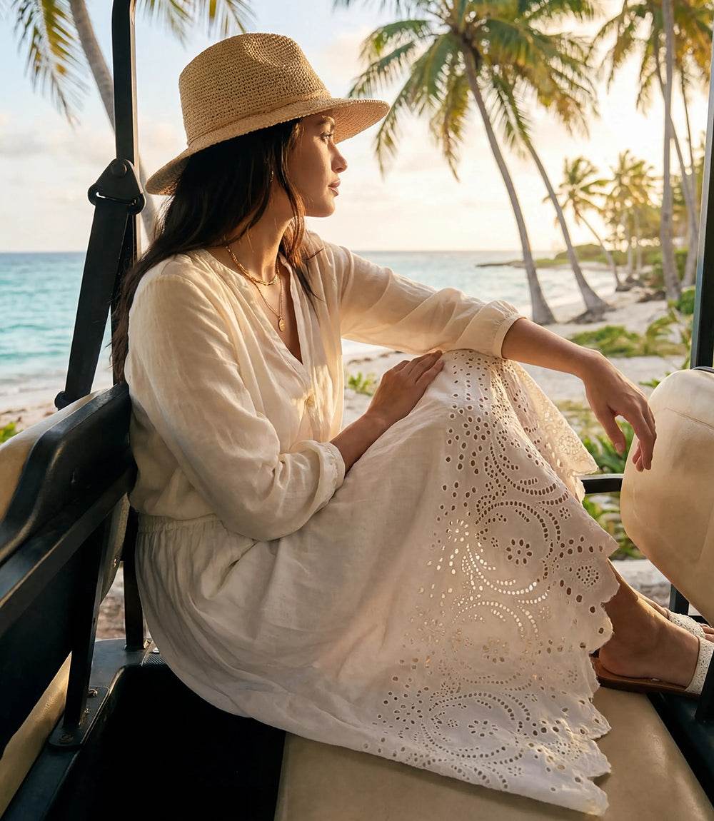 Woman in a white dress and straw hat sitting in a vehicle on a beach with palm trees and ocean in the background.