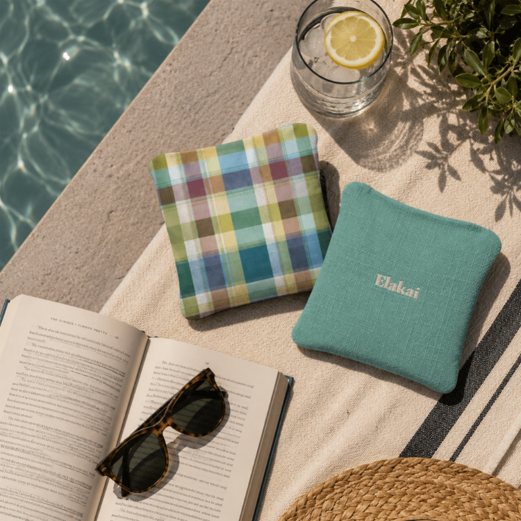 Two cushions, one plaid and one teal with 'Elakai' branding, next to an open book and sunglasses by a pool.