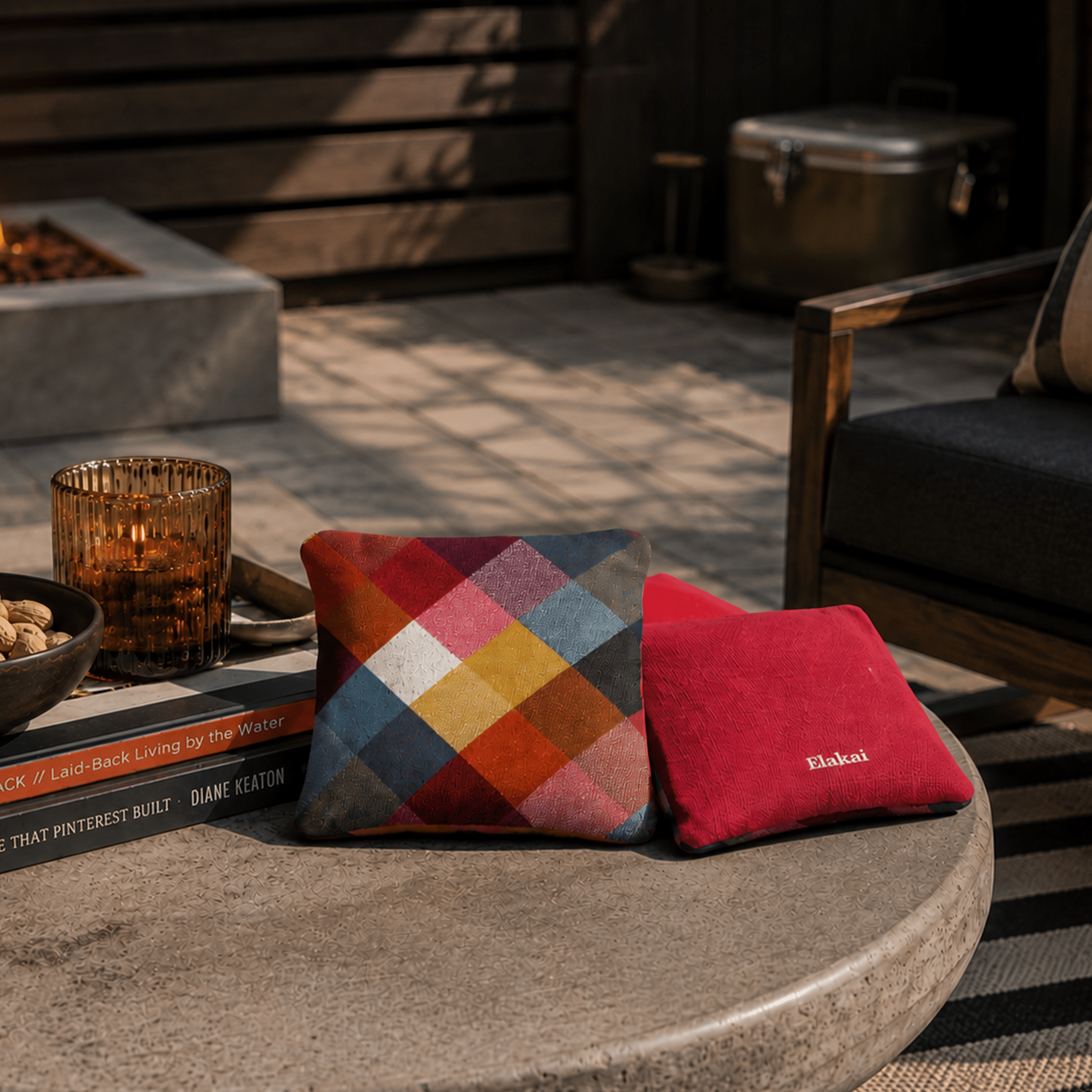 Two cushions, one multicolored and one red, on a stone surface with a fire pit and outdoor furniture in the background.