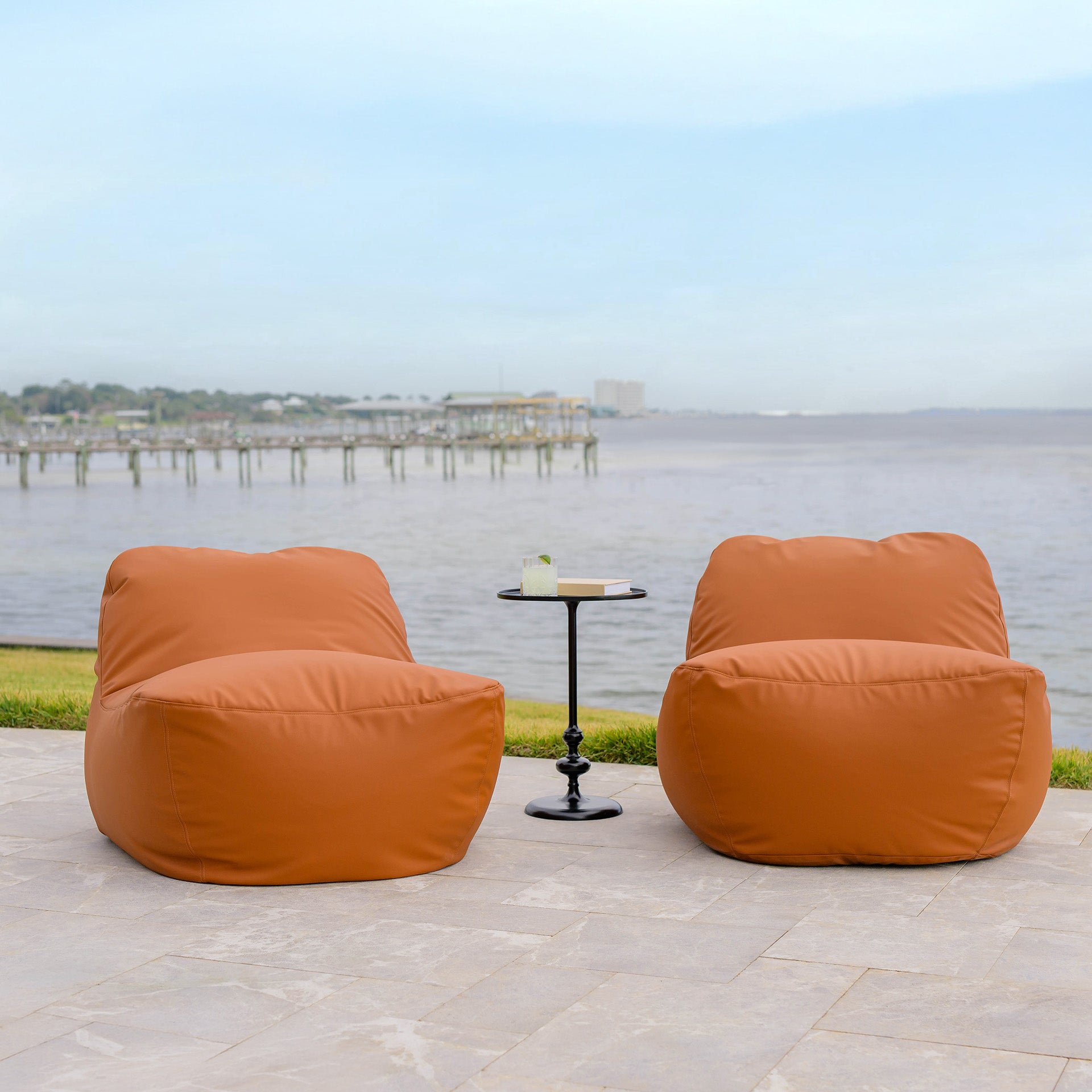 Two orange bean bags with a small table on a patio by a waterfront.