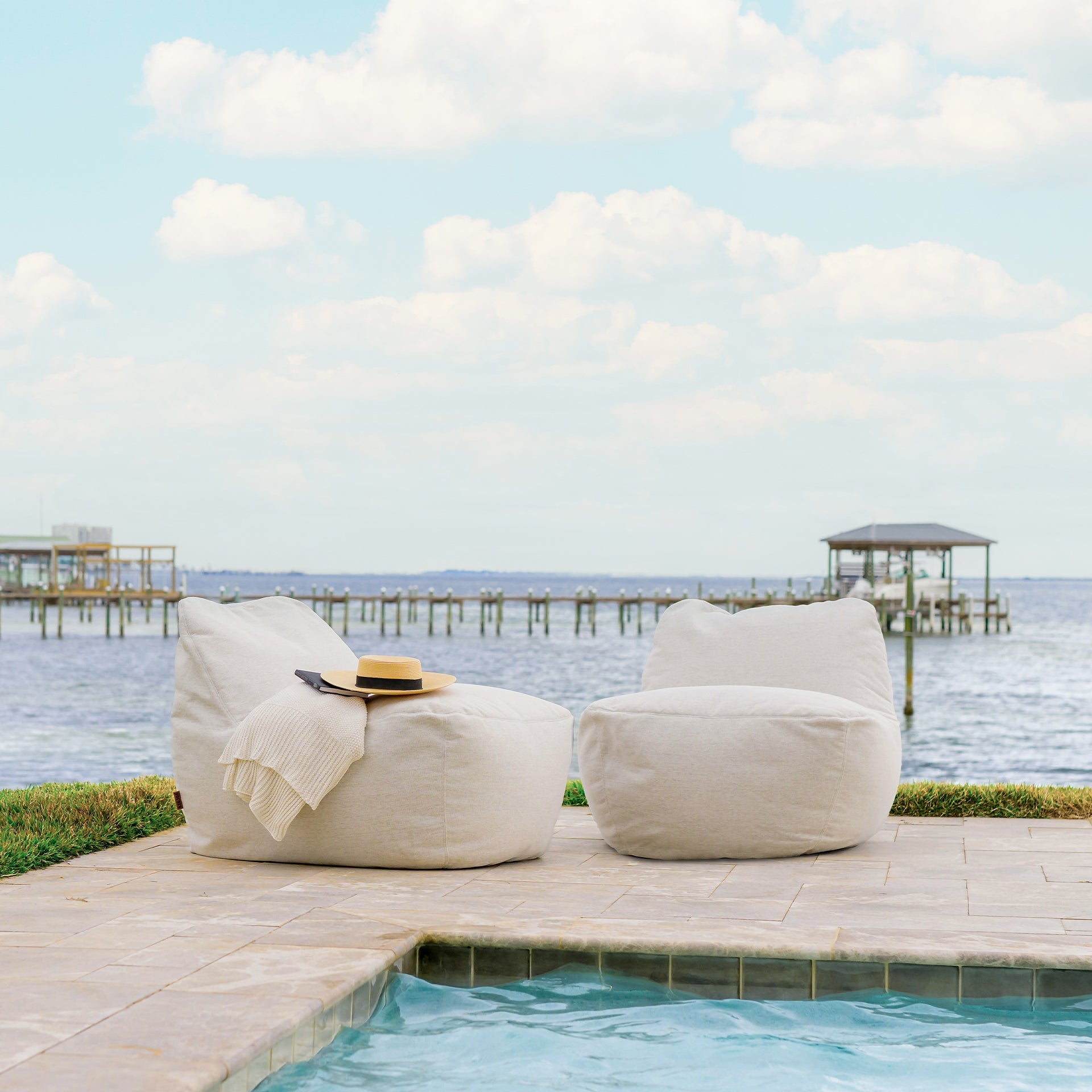 Two beige bean bags on a deck by a pool with a scenic view of water and sky.