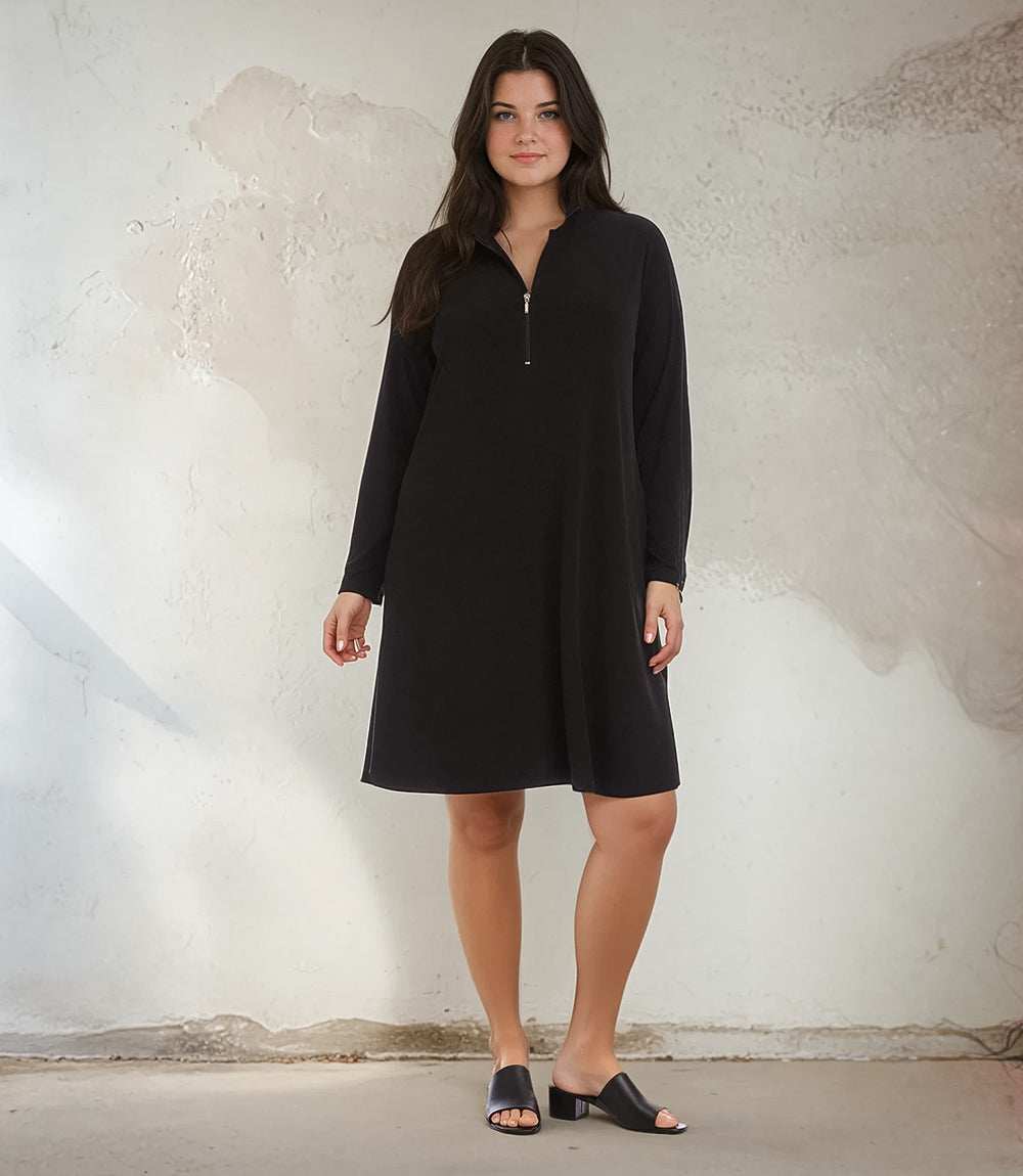 A woman modeling a black long-sleeve shift dress with a half-zip neckline, standing against a textured off-white wall.