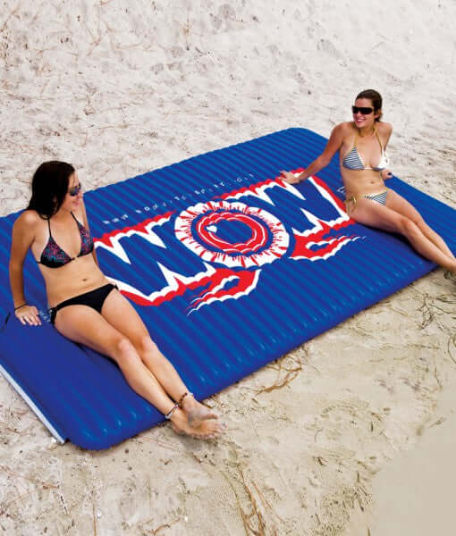 Two women sitting on a blue inflatable mat with 'WOW' branding on a sandy beach.