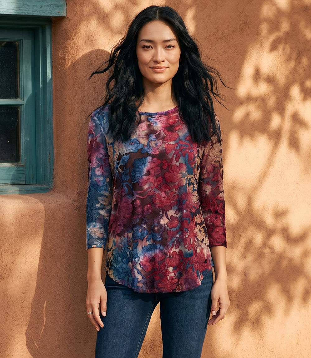 A woman wearing a three-quarter sleeve floral top with blue, red, and purple tones, paired with dark blue jeans, standing against a warm stucco wall with leaf shadows.