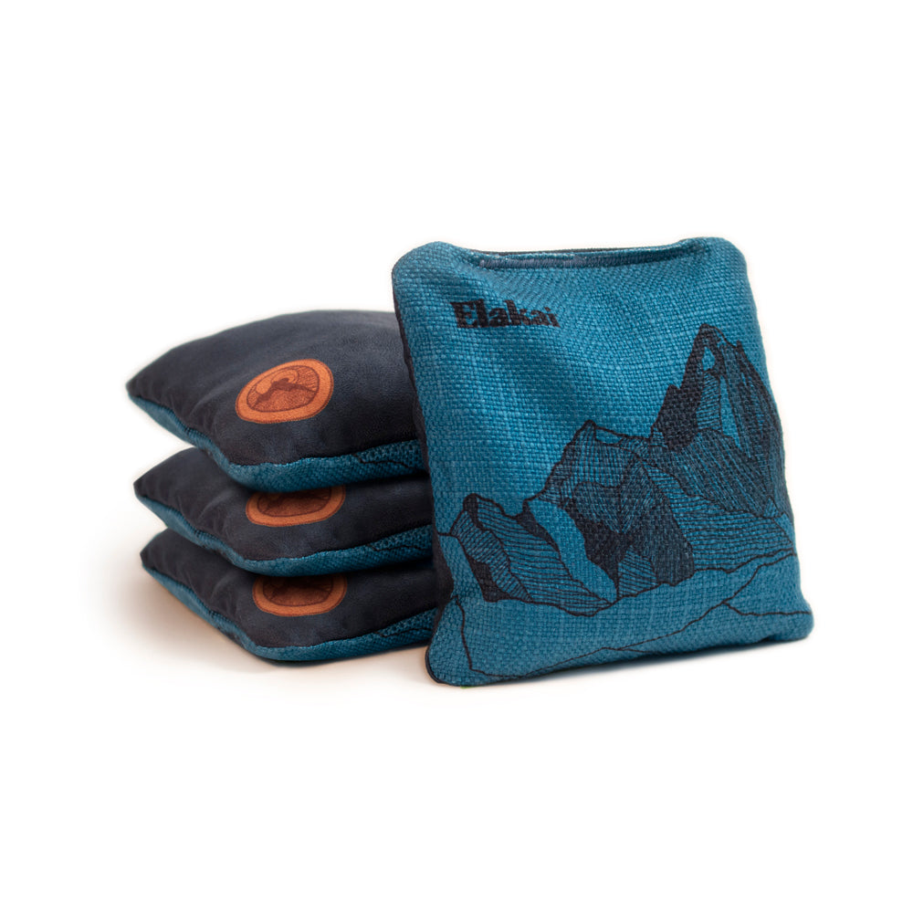 Three blue cornhole bags with a mountain design and one orange bag on a white background.