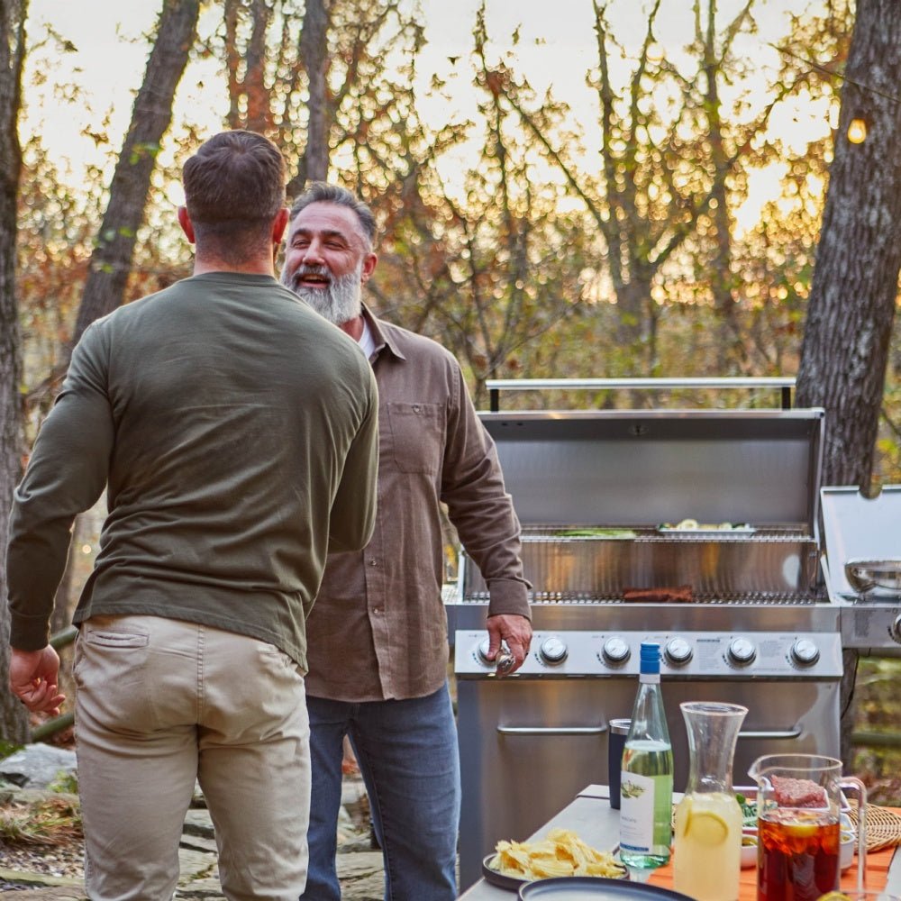 Two men standing outdoors by a grill with trees and sunset in the background
