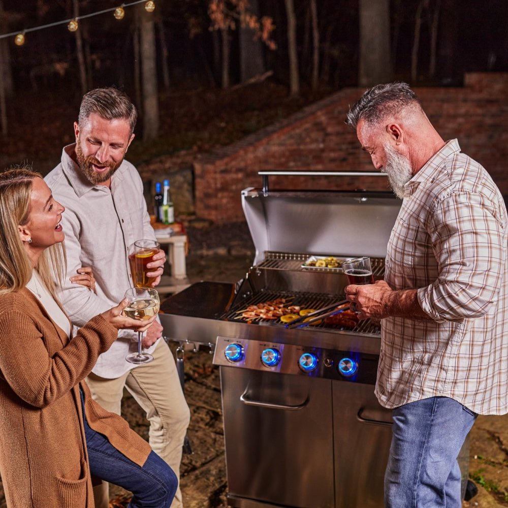 Three people around a grill with food cooking outdoors at night.