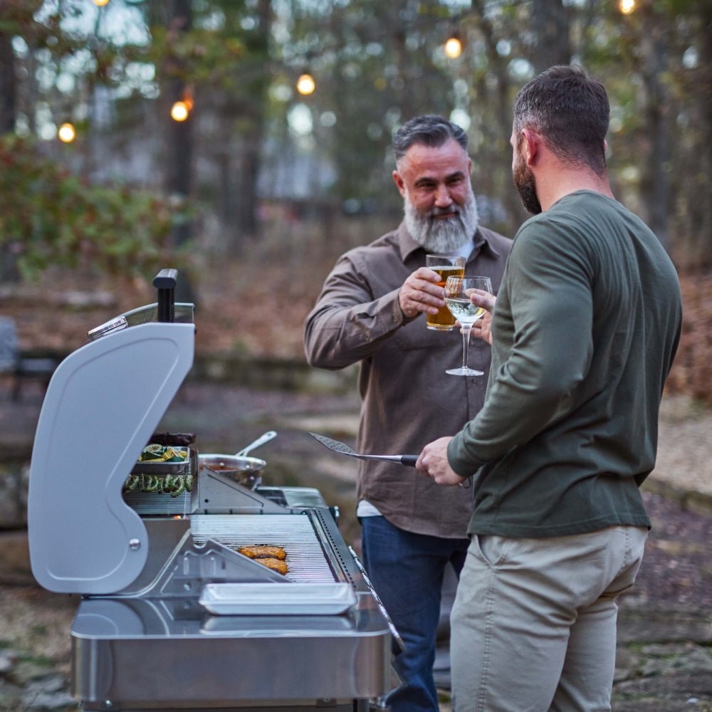 Two men grilling outdoors with a portable grill, one holding a beer.