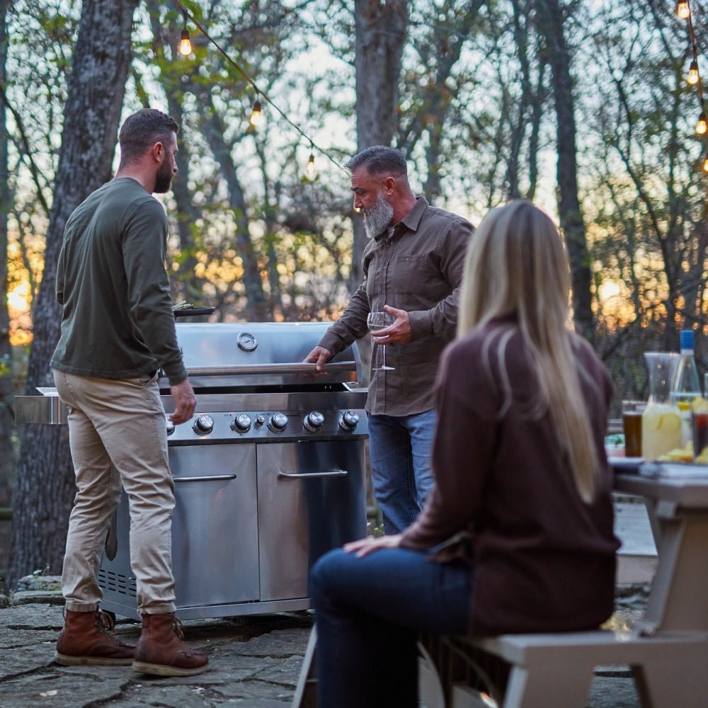 Three people around a grill in an outdoor setting with trees and a sunset.