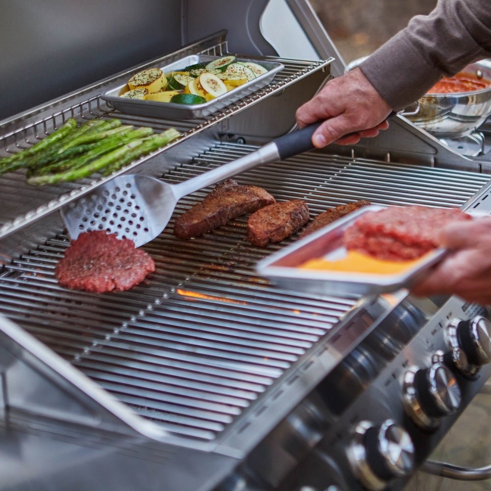 Person grilling vegetables and meat on a barbecue with a spatula.