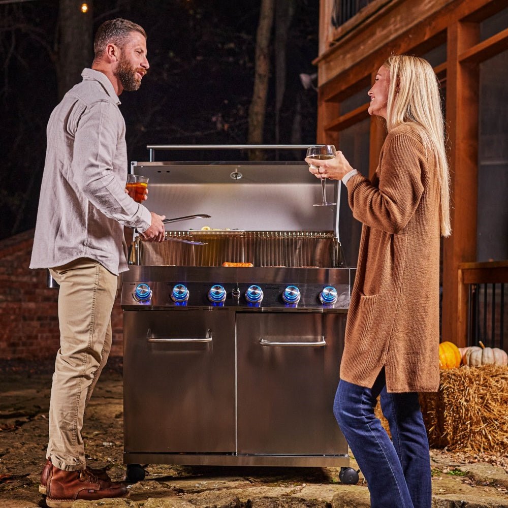 Two people standing next to a large outdoor grill in a rustic setting.